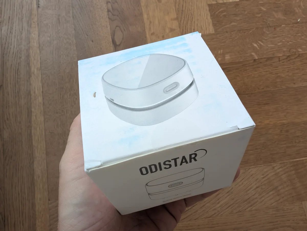 Stolní vysavač Odistar do ruky? Otestoval jsem za vás! 1 Bílá krabička stolního vysavače Odistar držená v ruce nad dřevěnou podlahou